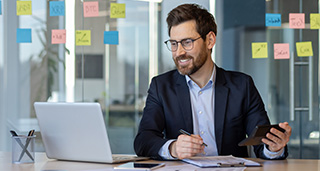 man in a suit working on a laptop while smiling holding a tablet with sticky notes on the wall related to business and productivity organized workspace 1